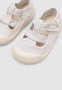 Baskets en toile beige clair avec un bout arrondi, deux sangles Velcro réglables et une semelle en caoutchouc texturé. Design minimaliste avec des découpes.