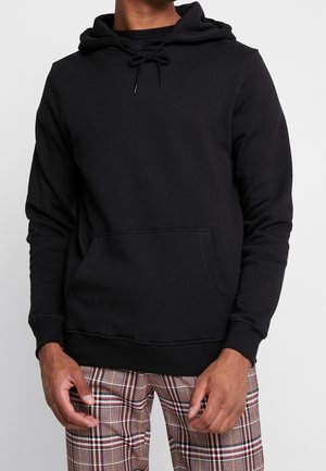 Man draagt een zwarte pullover hoodie met voorzak en veelkleurige geruite broek, staand tegen een effen witte achtergrond.