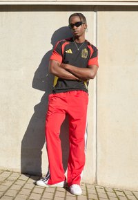 Sort og rød atletisk skjorte med gule accenter, røde bukser med sidelister og hvide sneakers. Stående mod en betonvæg.