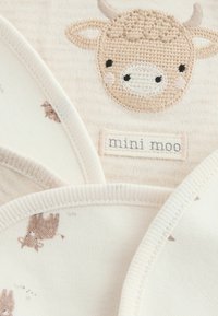 Smėlio spalvos medvilninis audinys, siuvinėta karvės veido aplikacija. Etiketėje parašyta „mini moo“. Audinys turi subtilius rudus gyvūnų raštus ir minkštą tekstūrą.