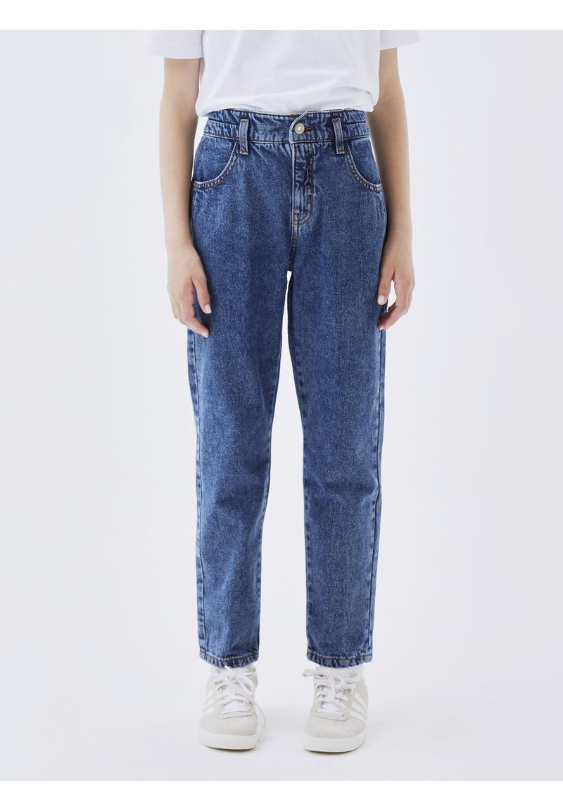Personne portant un jean en denim bleu taille haute, un t-shirt blanc et des baskets blanches, debout devant un fond uni.