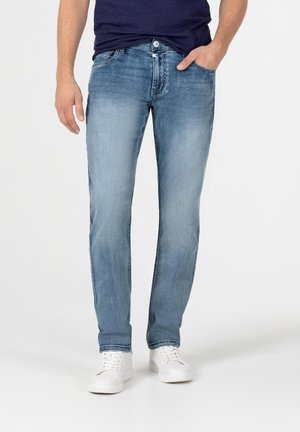 Man draagt lichtblauwe slim-fit jeans, een marineblauw overhemd en witte sneakers, staand met de rechterhand in de zak tegen een witte achtergrond.