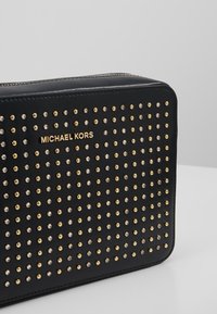 Clutch nera di Michael Kors con dettagli in borchie dorate e argentate e una cerniera dorata su uno sfondo grigio chiaro uniforme.