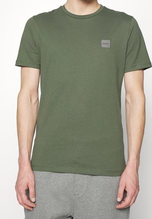 Mann trägt ein olivgrünes T-Shirt mit kurzen Ärmeln und einem kleinen "BOSS"-Aufnäher auf der Brust, kombiniert mit hellgrauen Hosen.