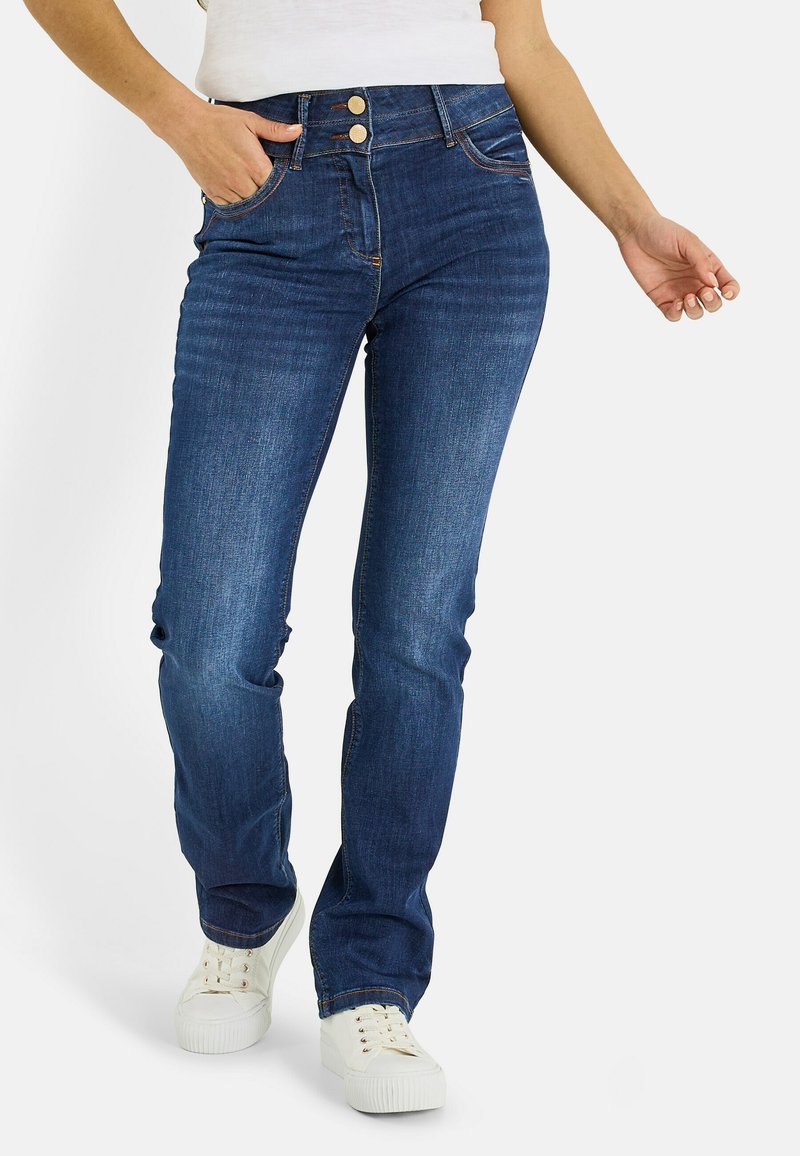 Vaqueros de mezclilla azul oscuro con corte ajustado, cintura alta, cierre de dos botones y bolsillos delanteros. Combinados con zapatillas blancas.