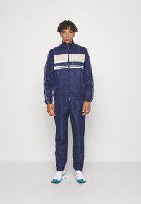Navy tracksuit with a beige and white stripe across the chest, featuring a zip-up jacket and matching pants. Smooth, sporty fabric.
