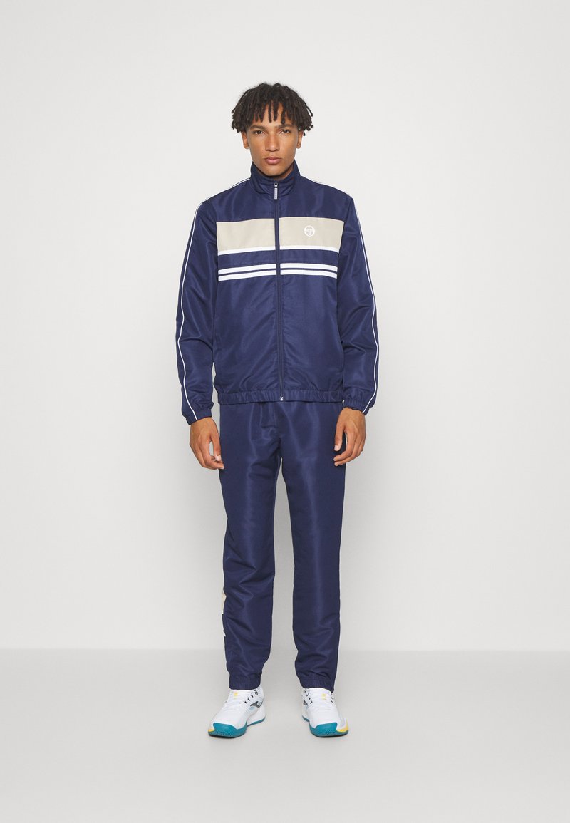 Navy tracksuit with a beige and white stripe across the chest, featuring a zip-up jacket and matching pants. Smooth, sporty fabric.
