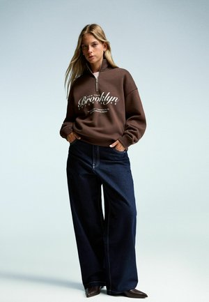 Brown zip-up sweatshirt with "Brooklyn" graphic, paired with wide-leg dark denim jeans featuring white stitching. Smooth fabric texture.