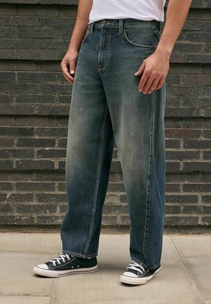 Mann trägt lockere dunkelblaue Jeans und schwarze Low-Top-Sneakers und steht auf dem Gehweg vor einer Ziegelsteinmauer.
