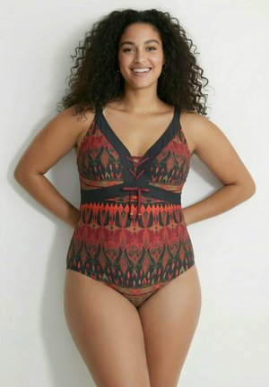 Donna sorridente con capelli ricci in piedi contro un muro bianco che indossa un costume intero rosso e nero a fantasia con dettaglio allacciato sul davanti.