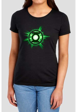 Frau trägt ein schwarzes T-Shirt mit grün leuchtendem Green Lantern-Logo und blaue Jeans, steht vor einfachem Hintergrund.