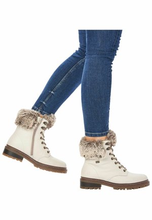 Personne portant un jean skinny bleu et des bottines blanches à lacets avec bordure en fourrure et fermeture éclair latérale, marchant sur un fond blanc.