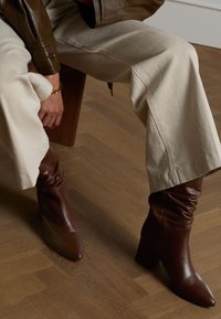 Bottes en cuir marron, arrivant au genou, avec un bout pointu et un design décontracté, posées sur un sol en bois à côté de pantalons larges de couleur claire.