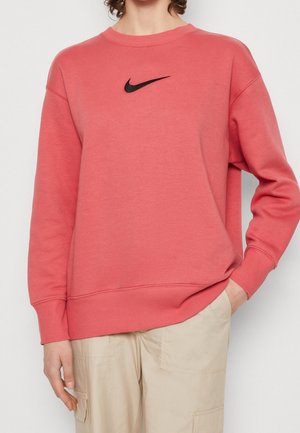 Personne portant un sweat-shirt Nike corail rose avec un logo swoosh noir et un pantalon cargo beige, main dans la poche.