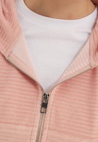 Haut de hoodies rose clair à fermeture éclair avec texture côtelée, détail de fermeture éclair argenté et un t-shirt blanc à col rond visible en dessous.