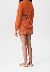 Orange krinklad kortärmad topp med lång ärm, knuten i ryggen. Matchande virkade minikjol med vågig fåll. Bruna snörskor.