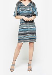 Robe à manches courtes en V avec un motif en zigzag bleu, noir et gris. Texture maille, longueur mi-cuisse, associée à des sandales à brides de cheville grises.