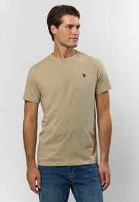 Beige bomullströja med kort ärm och rund hals, med en liten svart logotyp på vänster bröst. Bärs tillsammans med blå jeans.