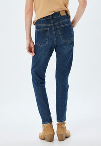 Jeans de mezclilla azul oscuro con forma ajustada, bolsillos traseros visibles y un ligero deshilachado en los dobladillos, combinados con botines de tobillo marrones.