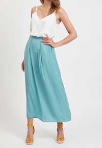 Frau in einer weißen ärmellosen Bluse, die in hoch taillierte, hellblaue weite Hosen gesteckt ist, mit felsigen, senfgelben Knöchelriemen-Heels.