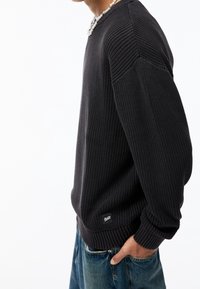 Suéter de punto en gris oscuro con textura acanalada, cuello redondo y hombros caídos. Presenta un pequeño logo en el dobladillo inferior y puños ajustados.