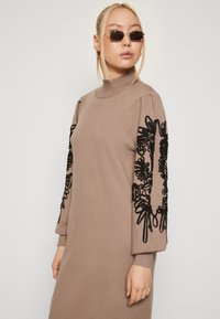 Strikkjole i beige med høj hals og lange ærmer. Udsmykket med detaljeret sort broderi på ærmerne. Blød tekstur, afslappet pasform.