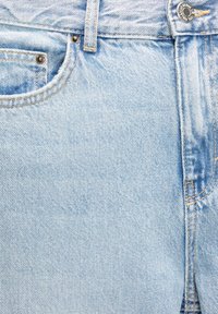 Détail avant d’un jean en denim bleu clair montrant un bouton métallique, une boucle de ceinture, une petite poche avant et des coutures visibles.