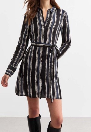 Kvinde iført en sort kjole med lange ærmer med lodrette hvide og blå striber, et bundet taljebånd og sorte knæhøje støvler.