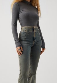 Grå ribbad långärmad topp i kombination med högmidjade, urblekta blåjeans med dragkedja fram och två metallknappar, hand vilande på sidan.