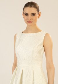 Robe blanche avec un motif de roses texturé, décolleté large et sans manches. Présente un corsage ajusté et une jupe plissée.