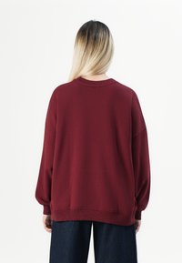 Person with long blonde ombre hair wearing a loose maroon sweater and dark wide-leg pants, shown from the back on a white background.