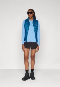Gilet rembourré bleu sur un haut à manches longues bleu clair, short noir et chaussures de sport sombres. Modèle portant des lunettes de soleil sombres, se tenant devant un fond uni.