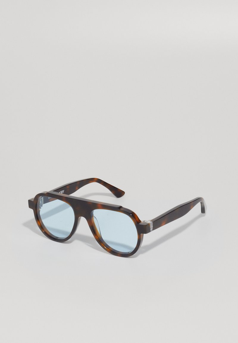 Γυαλιά ηλίου σε καβουροειδές σχέδιο με μπλε φακούς. Τετράγωνη, παχιά σχεδίαση με διαφανείς λεπτομέρειες κοντά στους μεντεσέδες. Λείος υφής, χωρίς ορατά λογότυπα.