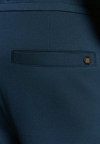 Diepblauwe stof met een gladde textuur, voorzien van een achterzak en een minimalistische zwart-gouden hexagonale accent.