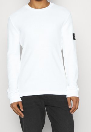 Hvid langærmet skjorte lavet af tekstureret stof med rund hals og ribbede manchetter, med et sort logo-patch på venstre ærme.