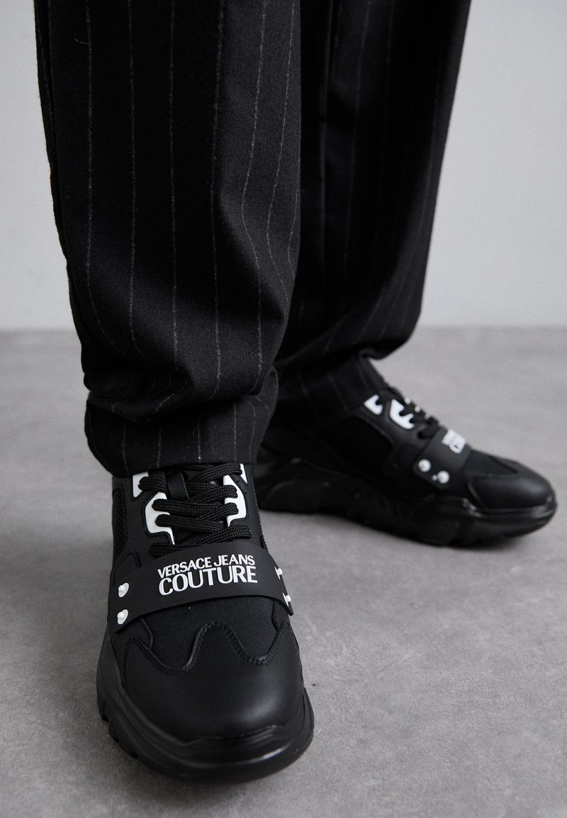 Schwarze Sneakers aus Netz- und Gummi-Materialien, ausgestattet mit weißen "VERSACE JEANS COUTURE"-Markierungen und silbernen Akzenten, kombiniert mit gestreiften Hosen.