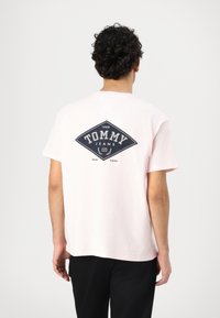 Camiseta de algodón rosa de manga corta, con un logo de diamante en navy en la parte posterior y texto que dice "TOMMY JEANS 1985 NEW YORK."
