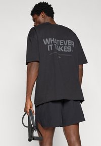 Čierne oversized tričko s nápisom „WHATEVER IT TAKES“ na chrbte, spárované s čiernymi športovými šortkami. Model drží fitness vybavenie.