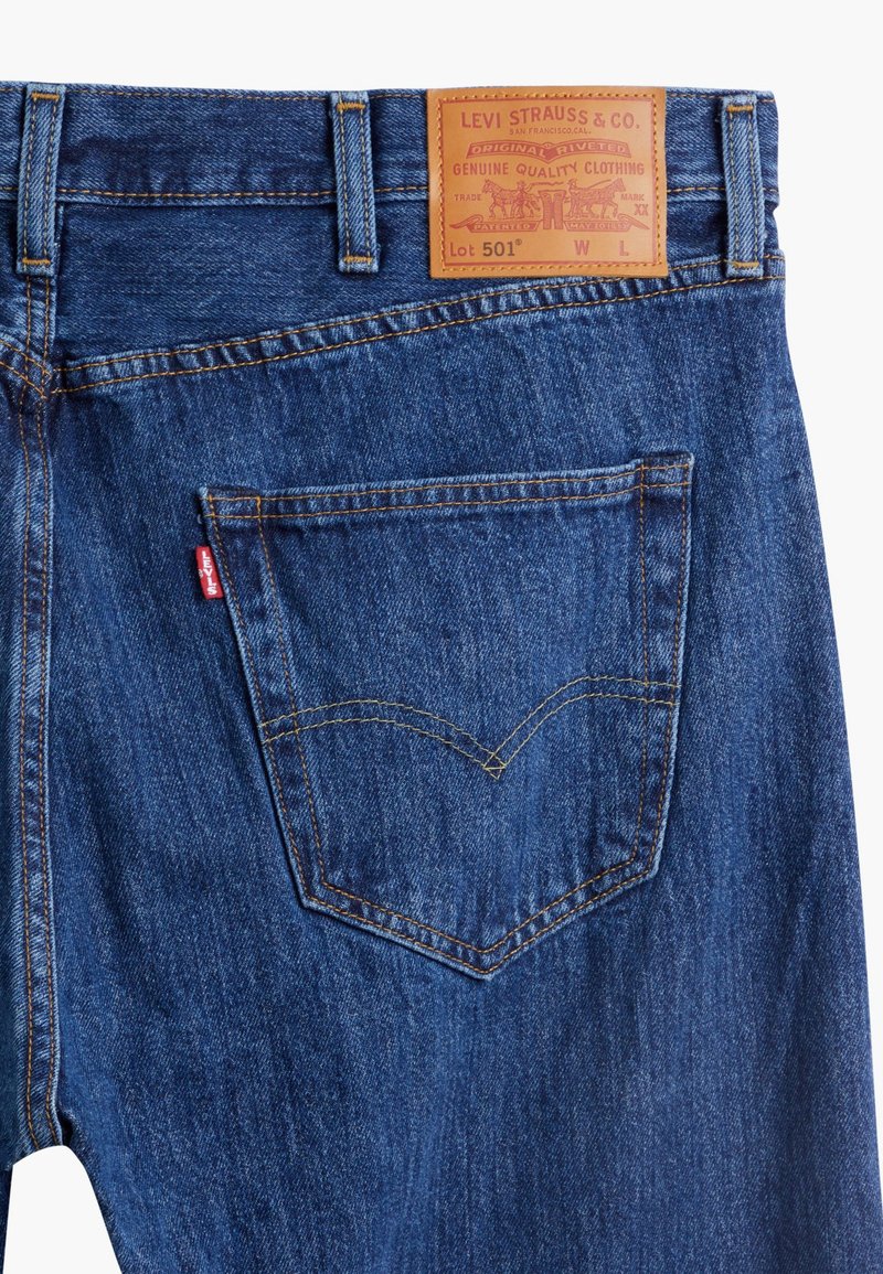 Jean en denim bleu avec une patch en cuir sur la taille affichant "Levi Strauss & Co." L'arrière présente un petit logo rouge et une couture de poche classique.