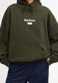 Personne portant un sweat à capuche Barbour vert olive avec une poche avant et une capuche à cordon, associé à un pantalon bleu foncé.