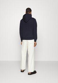 Felpa blu navy con cappuccio regolabile, vestibilità comoda. Abbinata a pantaloni bianchi larghi e calzature nere. Tessuto liscio, senza motivi visibili.