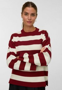 Pull rayé rouge et crème avec un col rond, des manches longues et des poignets côtelés. Le matériau semble doux avec une coupe décontractée et des fentes latérales.