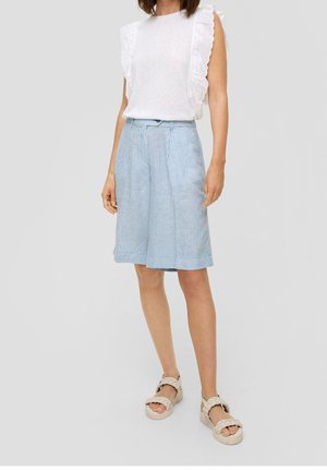 Vrouw draagt wit mouwloos topje met ruches, blauw-witte gestreepte knielange shorts en beige gevlochten sandalen, staand op een grijze achtergrond.