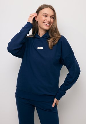 Uśmiechnięta kobieta z długimi włosami, ubrana w granatową bluzę i spodnie, dotyka włosów jedną ręką, stoi na tle jednolitego jasnego tła.