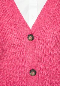 Rosa stickad kofta med djup V-ringning, med två svarta knappar framtill och ett texturerat tygmönster.