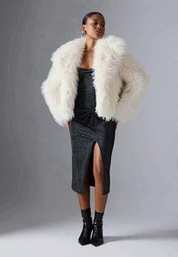 White faux fur coat over a black sparkly dress with a thigh slit, paired with black heeled boots. Simple background.