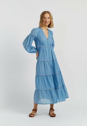 Glunderende vrouw in een lange blauwe laagjesjurk met knopen en pofmouwen, staand met één hand op haar heup en bruine sandalen.