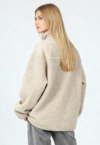 Giacca in pile beige con colletto alto e corpo strutturato, dotata di area spalle in tessuto liscio e vestibilità rilassata.