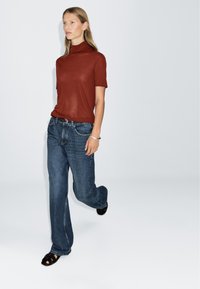 Top in turtleneck a maniche corte color ruggine, abbinato a jeans in denim blu con vestibilità ampia e sandali neri con lacci, in posa mentre cammina.