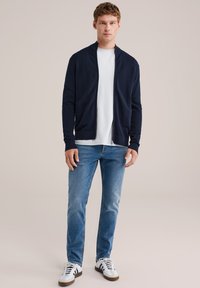 Cárdigan navy con cremallera sobre una camisa blanca, combinado con jeans azul claro y zapatillas blancas. El cárdigan presenta un corte relajado y una tela texturizada.